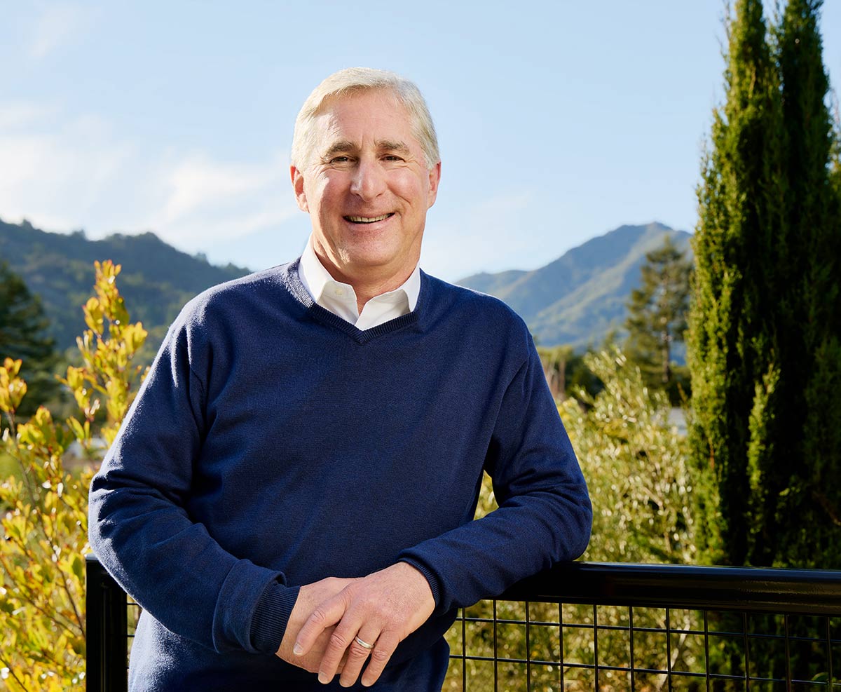 Jerry Kurland leaning against a railing, smiling, with a mountain vista behind him