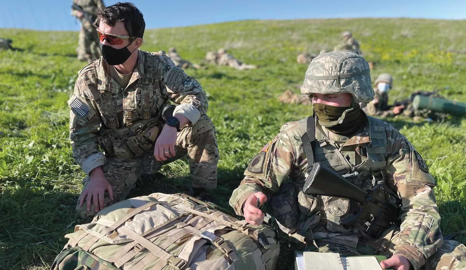 two ROTC cadets wearing full camouflage and sittin on a grassy hill