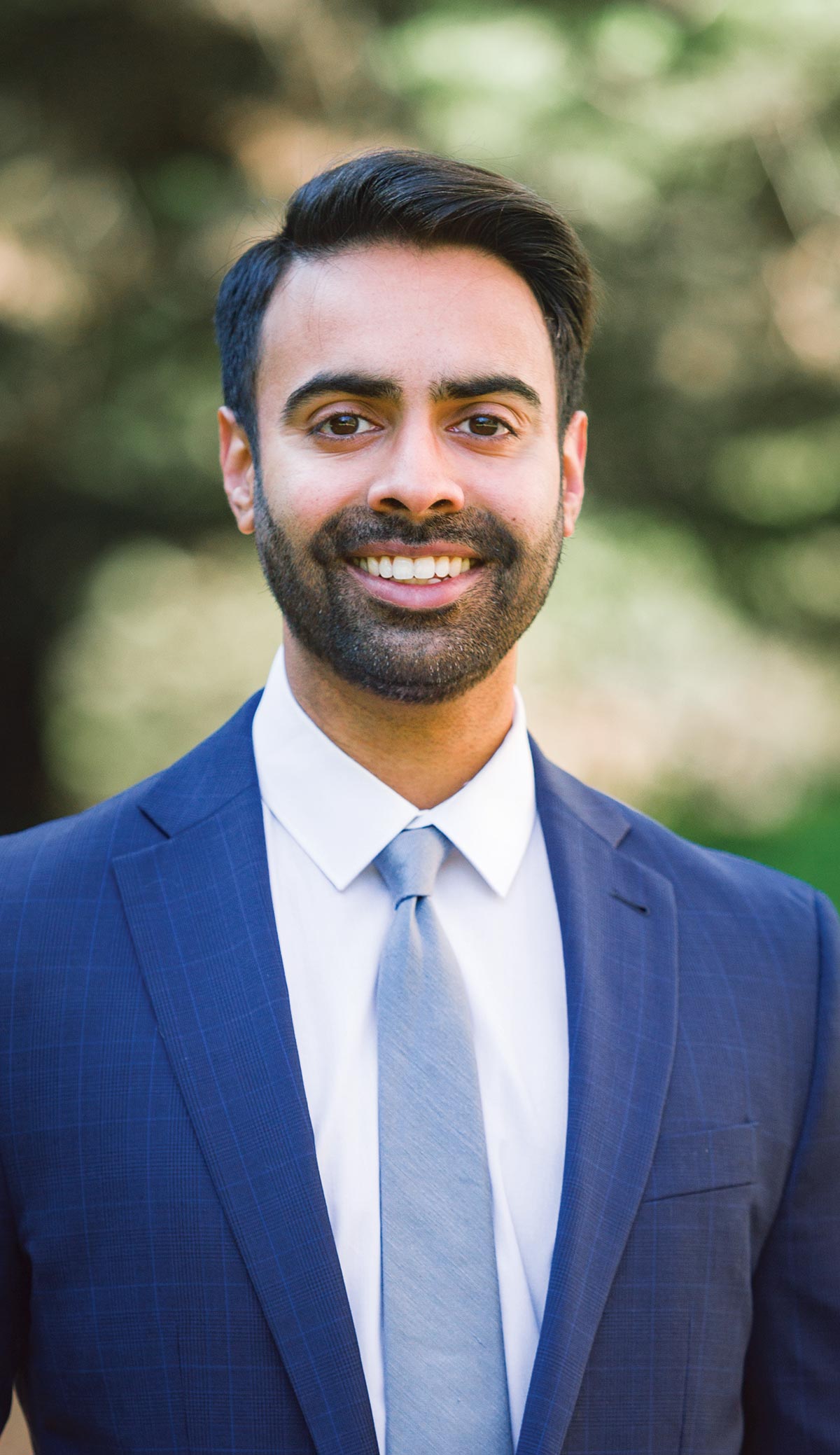 A portrait photograph of Nikesh Patel smiling as he poses for a picture in his business attire outside