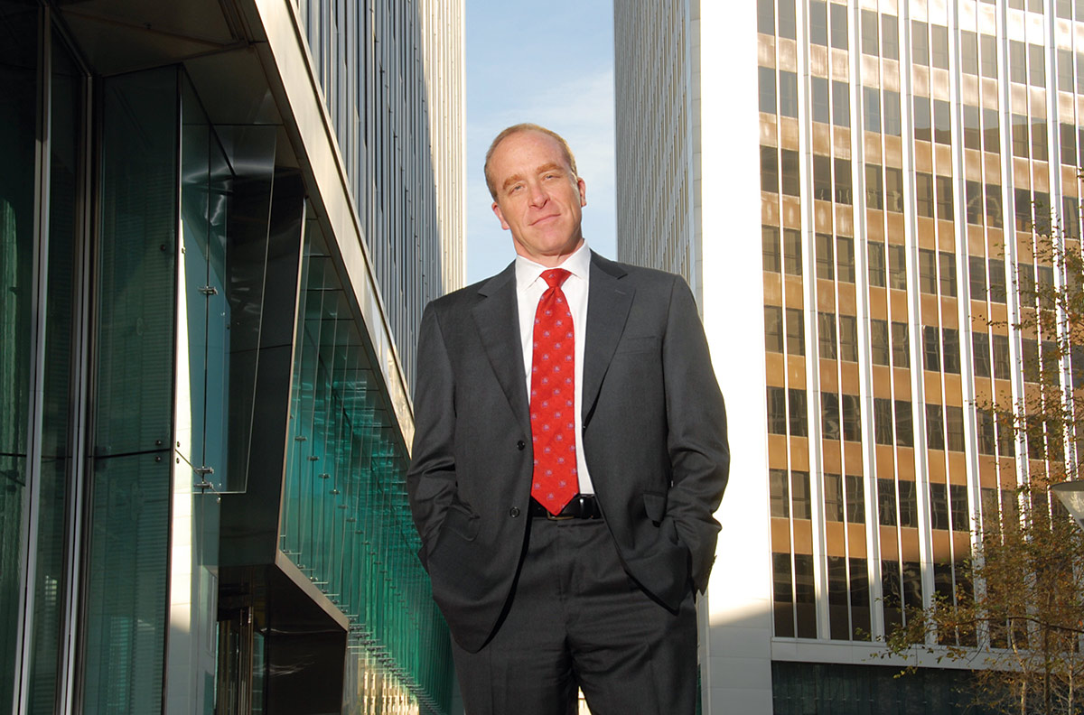 A landscape photograph of Scott Edelman grinning while he poses for a picture in his business attire outside in between buildings