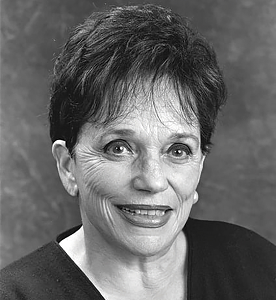 A black and white portrait headshot photograph of Babette Barton smiling in white earrings and a black shirt