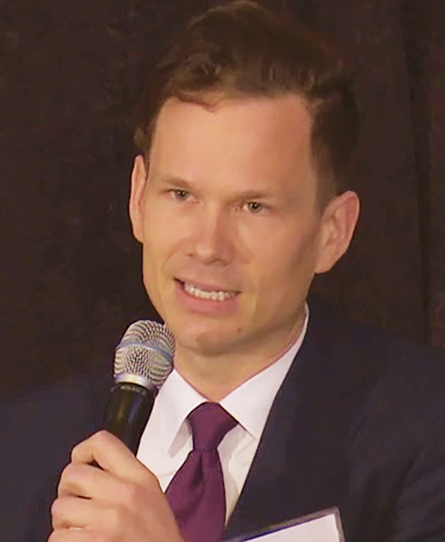 A portrait headshot photograph of Dan Mistak talking into a black microphone in a dark violet tie