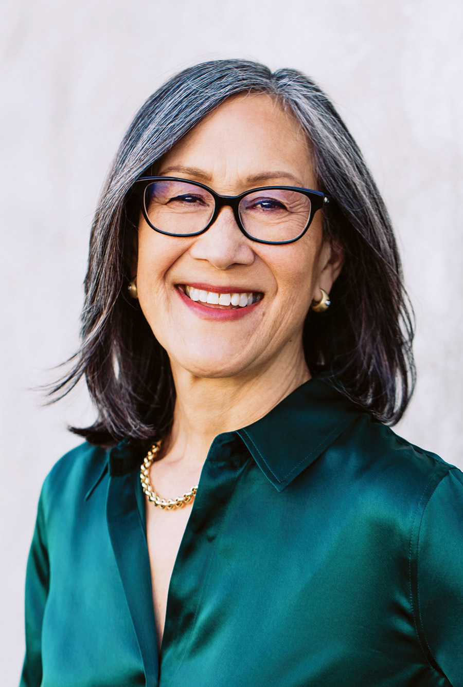 A portrait headshot photograph of Kathryn Ma smiling in an emerald green silk material button-up dress shirt with a golden necklace and see through black outer frame color glasses plus two golden earrings