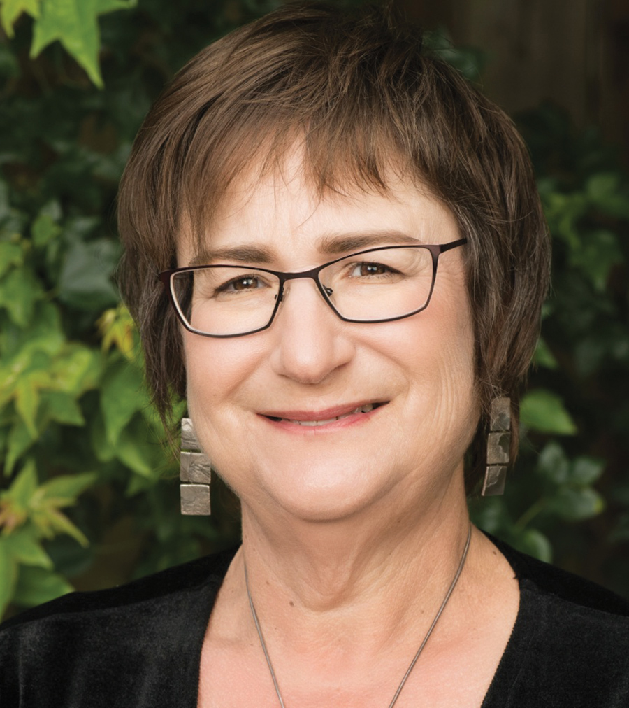 A portrait headshot photograph of Lauren Edelman smiling in a black shirt and gray necklace with grey earrings plus black outer frame see through glasses outside near some green leaves that are apart of small trees/shrubs