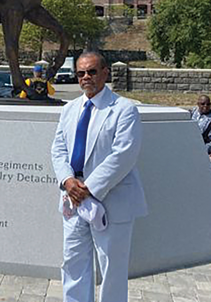 A portrait photograph of Leroy Wilson Jr. grinning in a light baby blue suit and dark blue tie plus black tinted colored sunglasses holding a hat in his hands as he poses for a picture in front of a Buffalo Soldier monument sculpture display outside
