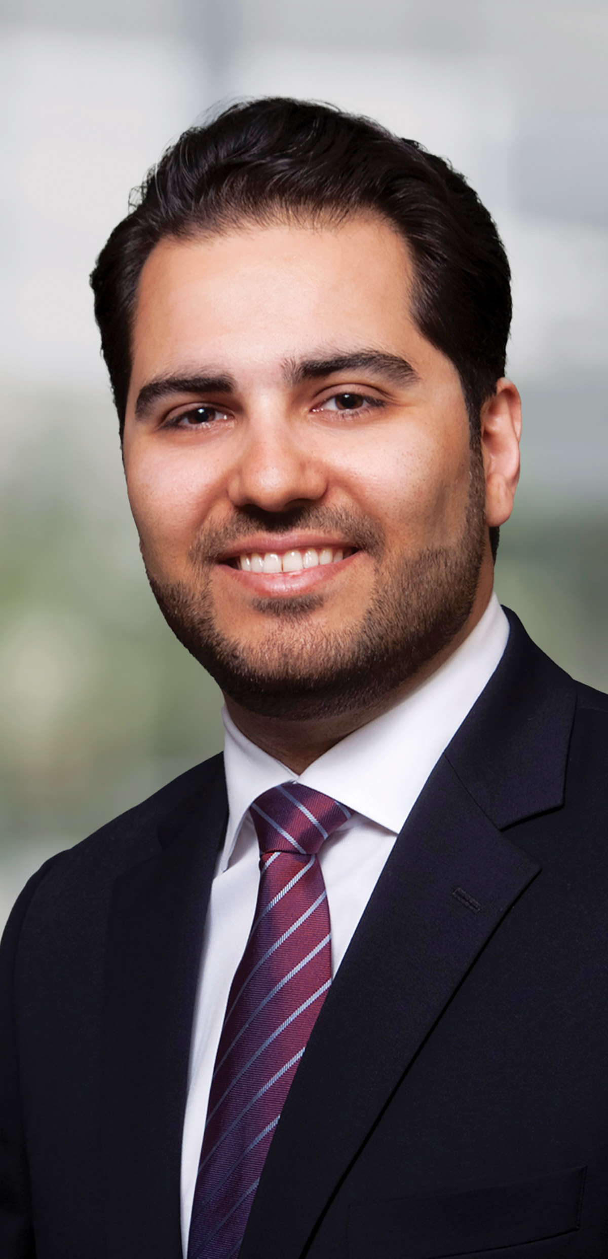A portrait headshot photograph of Mehdi Ansari smiling in a dark black suit and white button-up dress shirt underneath with a multi-colored tie (burgundy as the primary base color and gray lined stripes downward pattern as the secondary color)