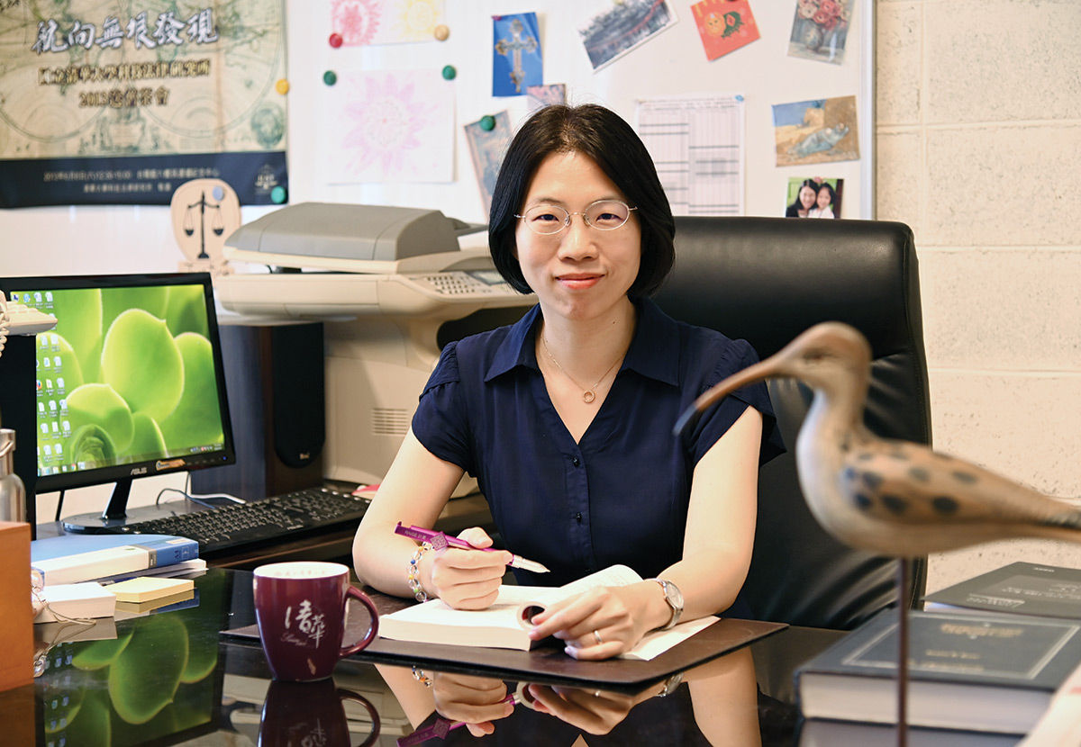 A landscape photograph of Yun-Hsien Diana Lin grinning in a dark navy blue button-up dress shirt and circular design necklace plus white outer frame colored glasses holding a violet pen and a white book sitting at her office desk nearby a coffee mug, computer, printer, table, bulletin white board, and a small bird ornament sculpture stand display at National Tsing Hua University