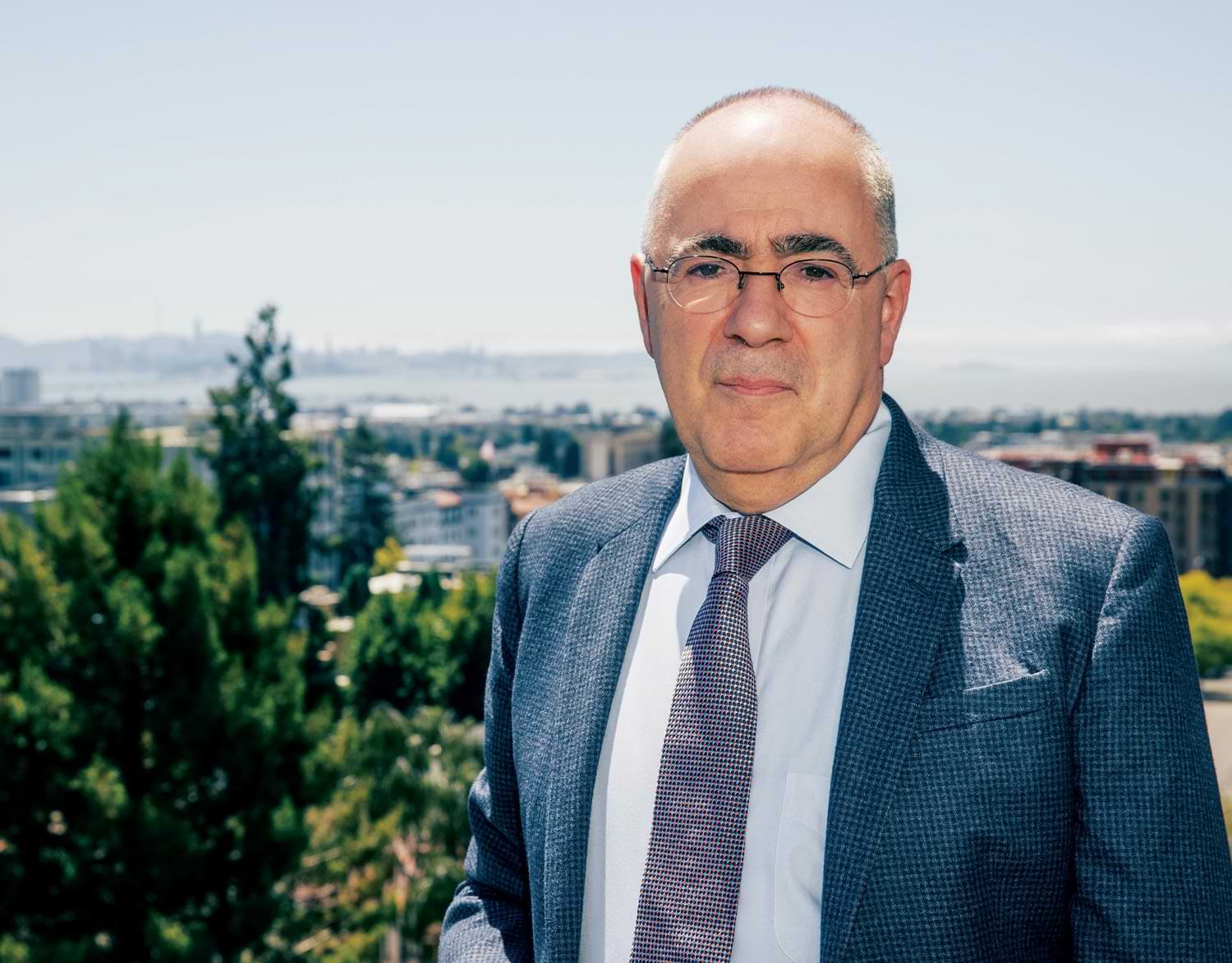 Hanoch Dagan photographed wearing a suit jacket, tie and thin rimmed glasses while standing against a city landscape on a bright day