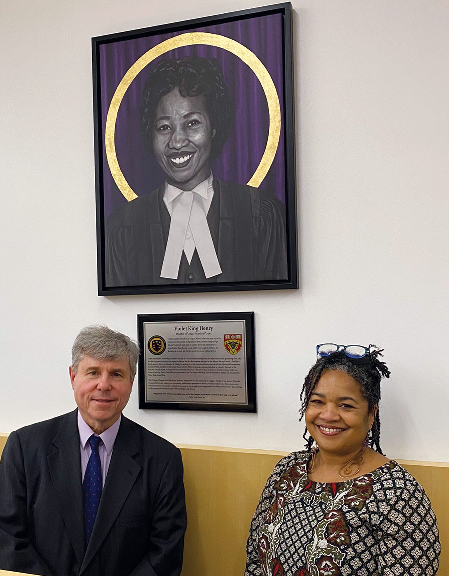 Portrait headshot photograph of Jo-Anne Henry (located on the right) smiling in a multi-color/floral pattern design dress top and prescription see through glasses on top of her head sitting down next to a man in a black suit and dark blue tie/light violet button-up dress shirt as they are both sitting next to each other below a portrait of Violet King Henry