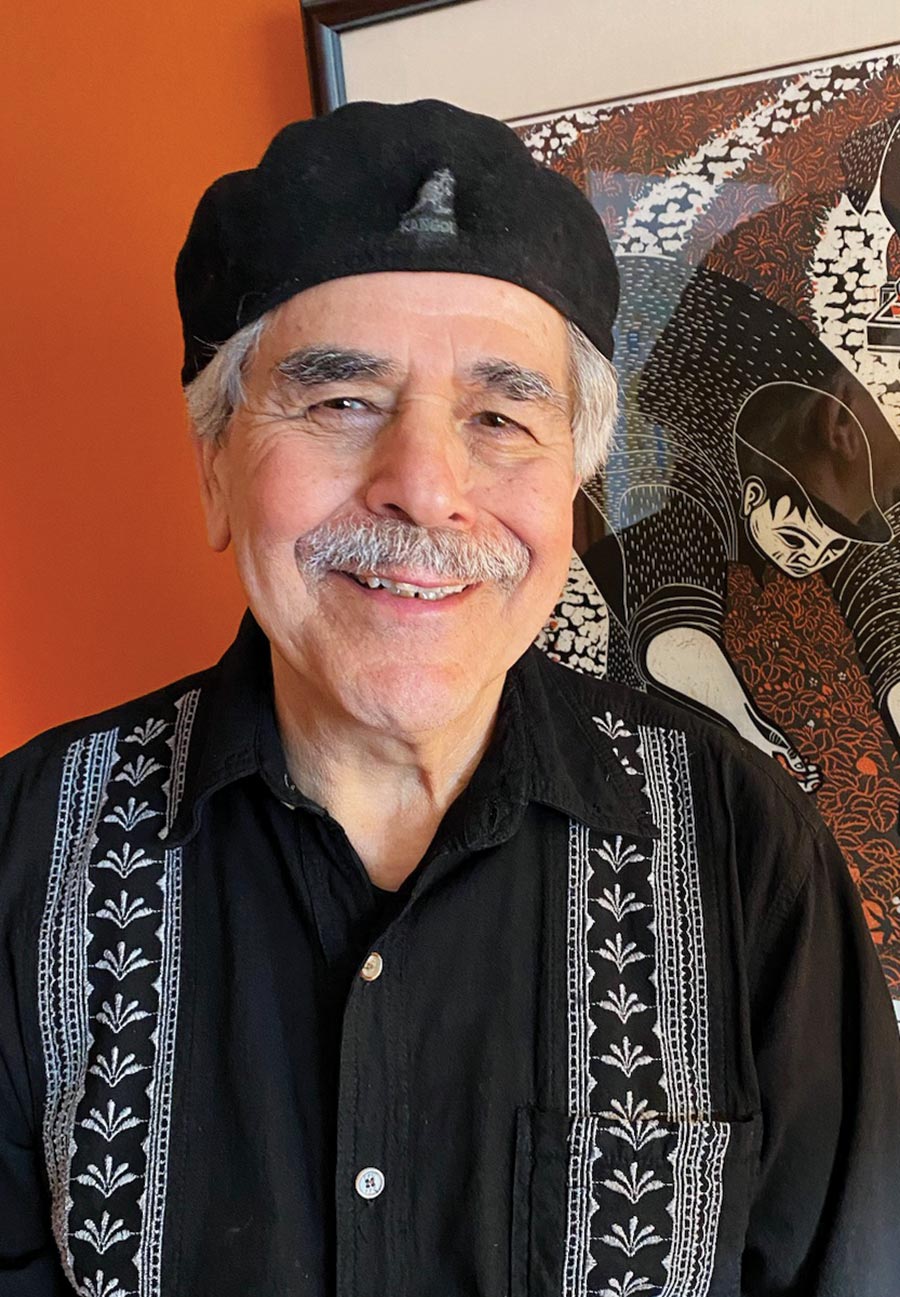 Portrait headshot photograph of José Padilla smiling in a black button-up dress shirt with white decorative plant/ribbon embroidered style pattern and a black hat as he poses for a picture nearby a minimalist picture frame design