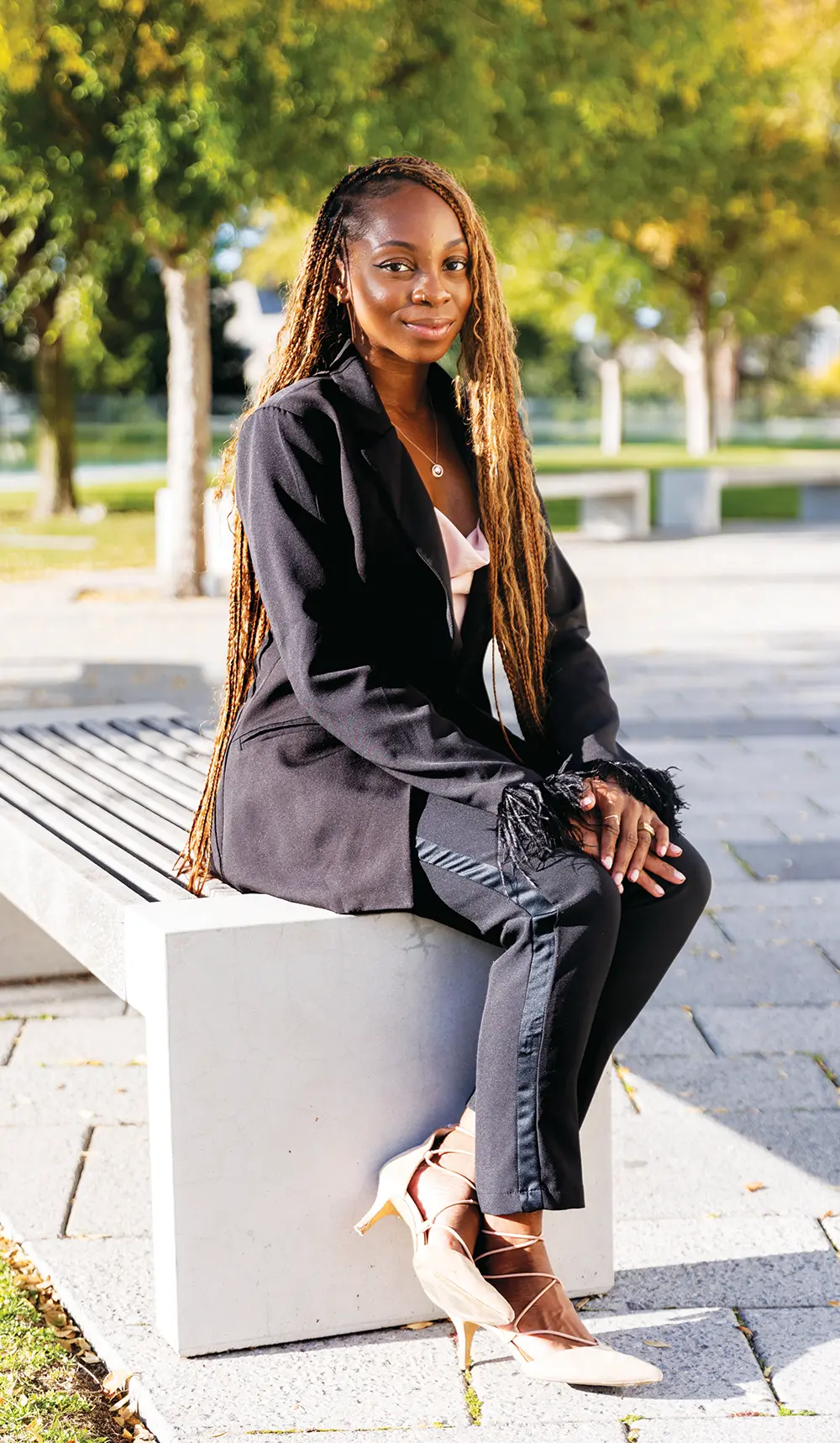Portrait close-up photograph view of Dayo Ajanaku grinning in a black open coat business blazer, pink blouse, necklace, black business dress pants, and beige high heel shoes as she is seated outside on a light grey bench somewhere during the day with her hands posed over each other on her knees