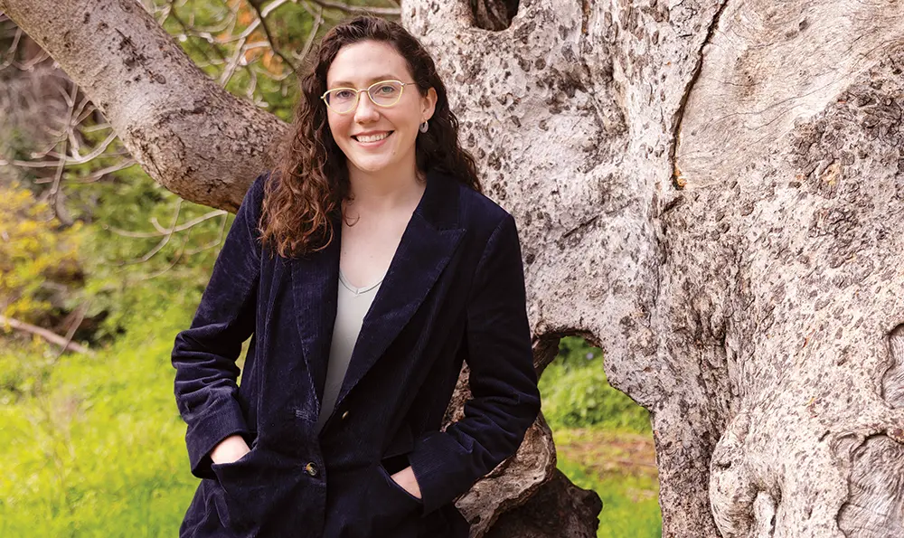Landscape close-up photograph view of Emma Lewis smiling in yellow outer frame see through prescription glasses, chrome-colored earrings, dark navy blue business overcoat, and a light grey blouse as she has her hands resting inside the overcoat's pockets while she poses/standing up against the side of a tree branch area outside somewhere