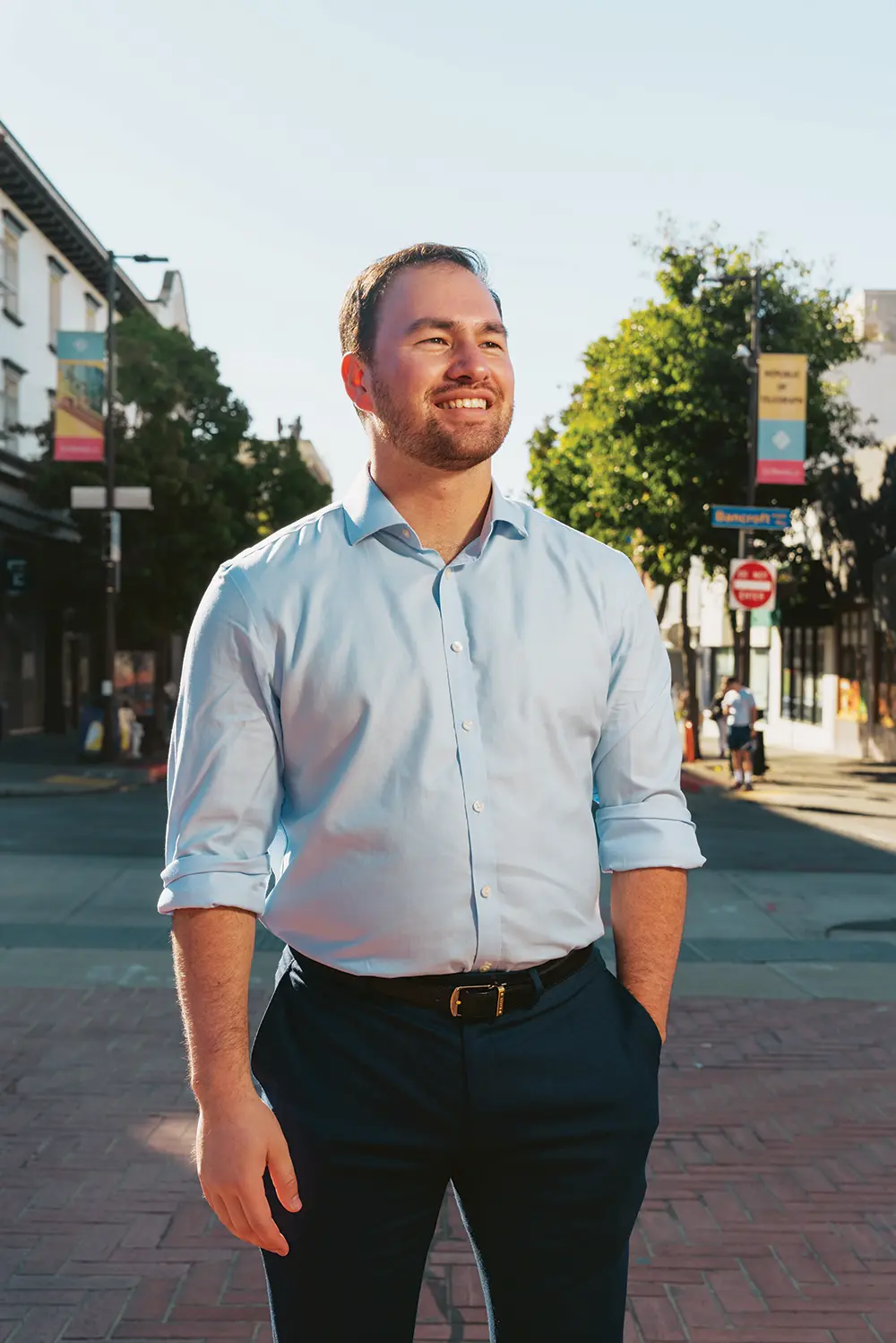 Portrait close-up photograph view of Josh Cayetano smiling in a light sky blue button-up dress shirt and dark navy blue business dress pants as he has his left arm resting inside his left pant pocket while the other right arm is freely resting in the air while he stands somewhere out in town at an intersection area during the day