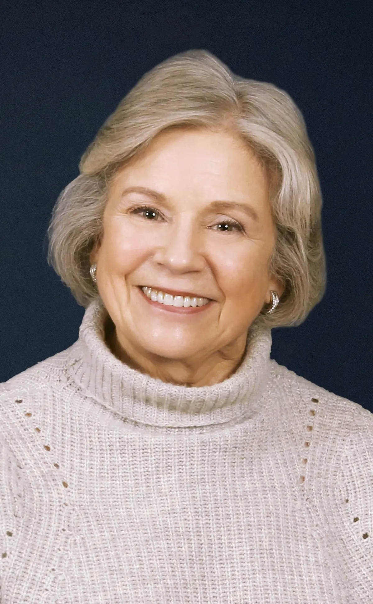 Portrait headshot close-up photograph view of Judith Droz Keyes smiling in a dark beige/cream colored turtleneck cardigan top and has on silver colored small circular earrings