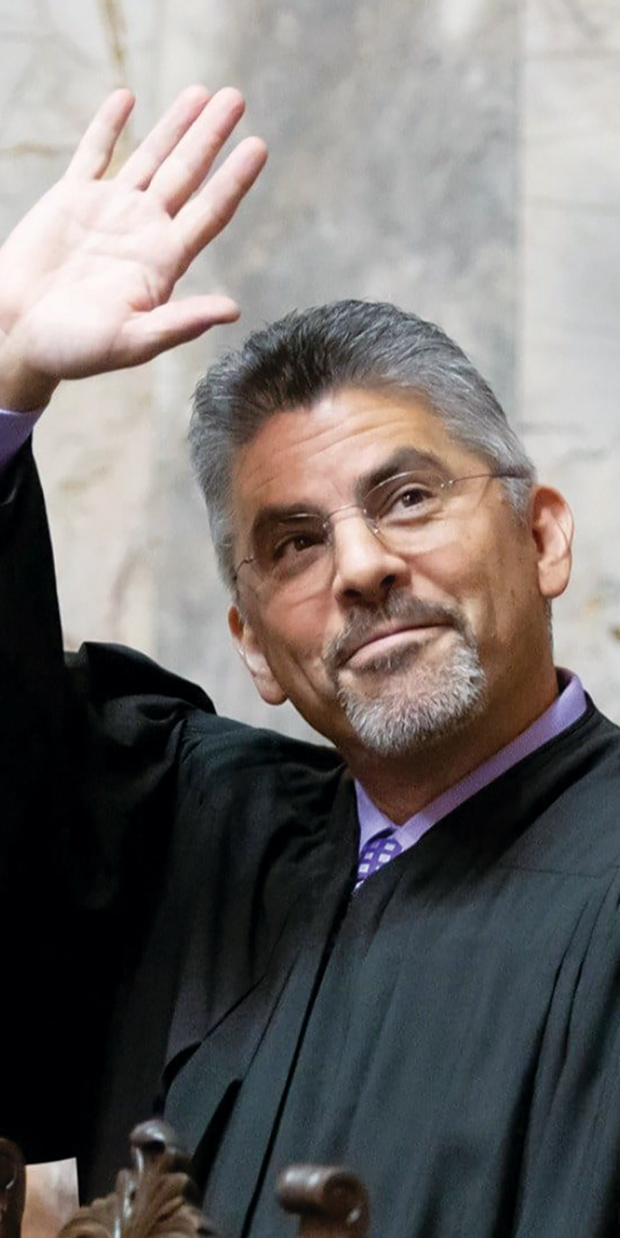 Portrait headshot close-up photograph view of Steven González grinning as he is in a black chief justice state supreme court gown with a light bright violet button-up dress shirt underneath with an equipped darker violet square shaped pattern tie while waving his right hand towards to what appears like an audience of some sort