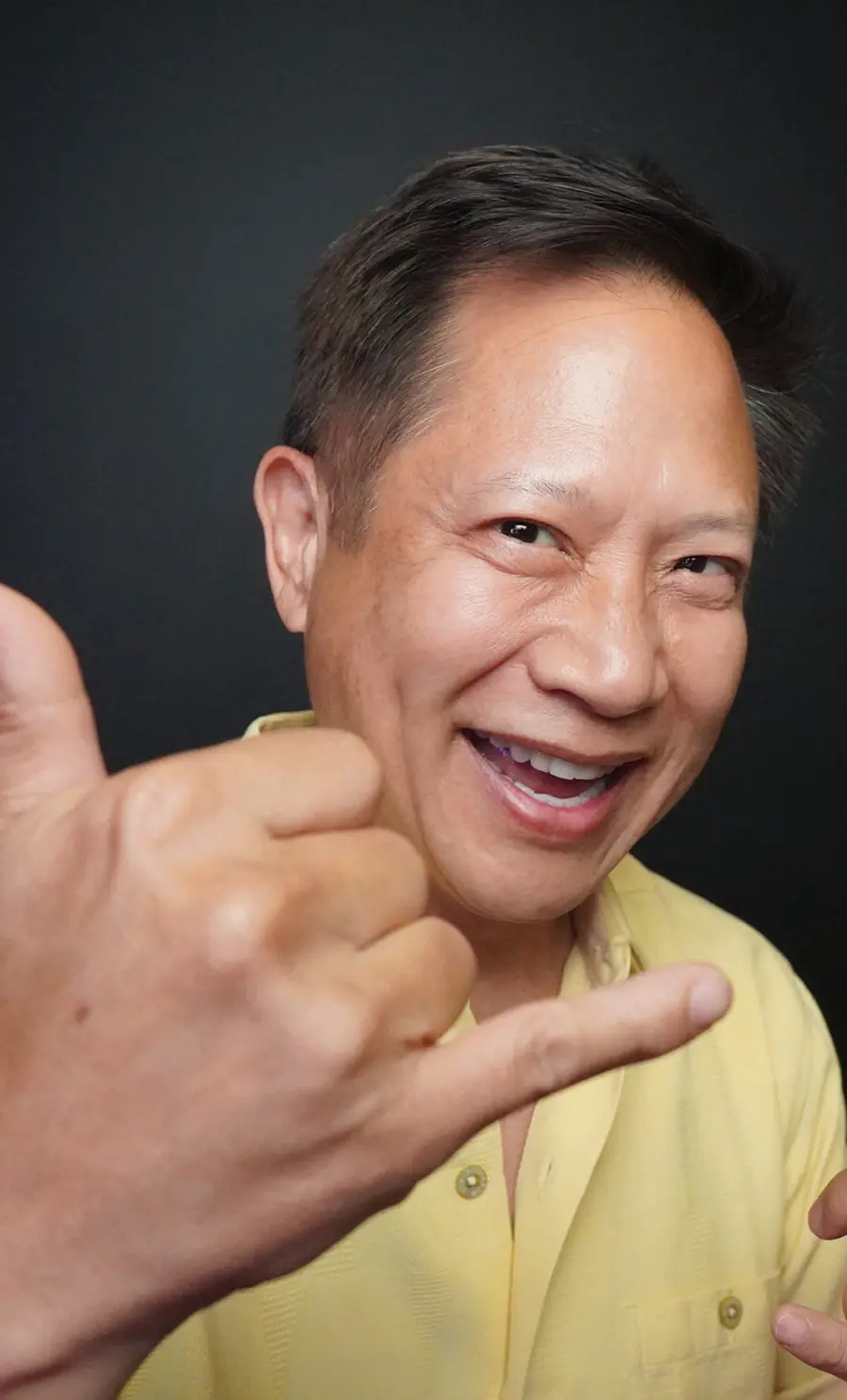 Portrait close-up photograph headshot view of Steve Sue smiling in a yellow button-up dress shirt as he has his right hand formed into a hang loose hand gesture sign manner