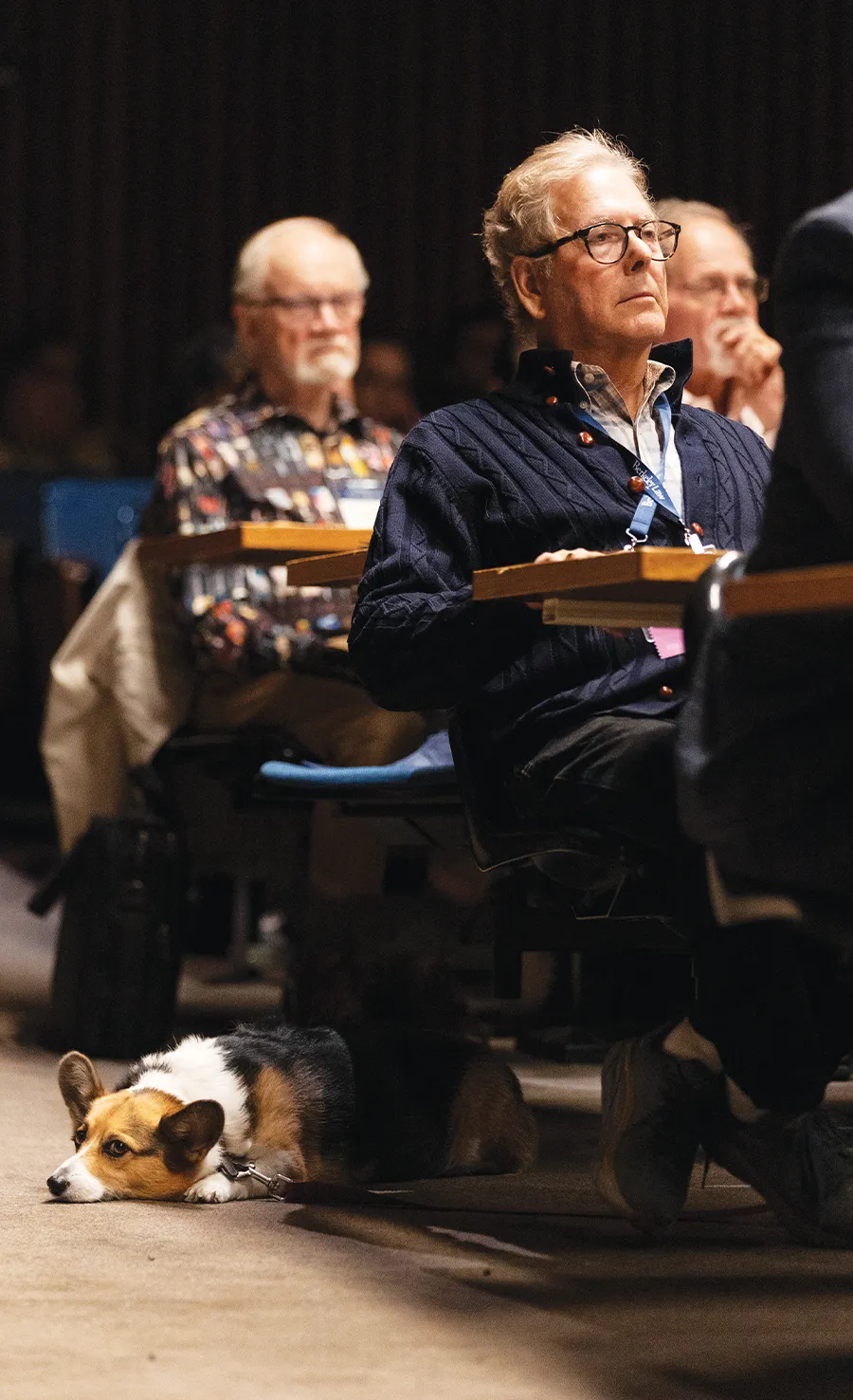 Jim Richman sitting in an auditorium with his dog lying on the floor beside him