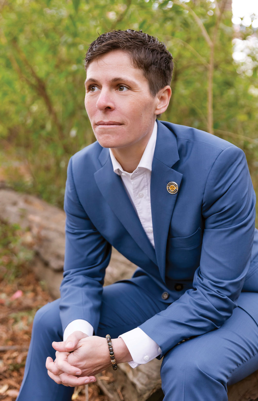 Person in a blue suit sitting on a rock with greenery in the background.