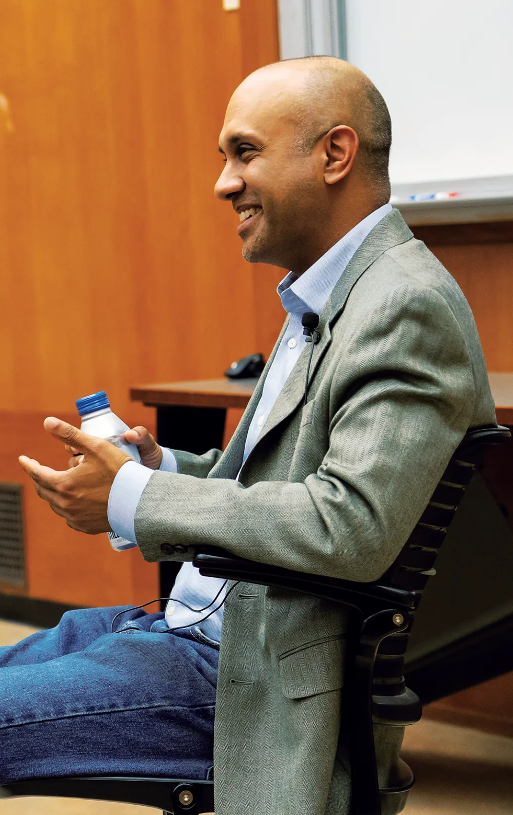 Man in a gray blazer sitting in a chair, holding a water bottle.