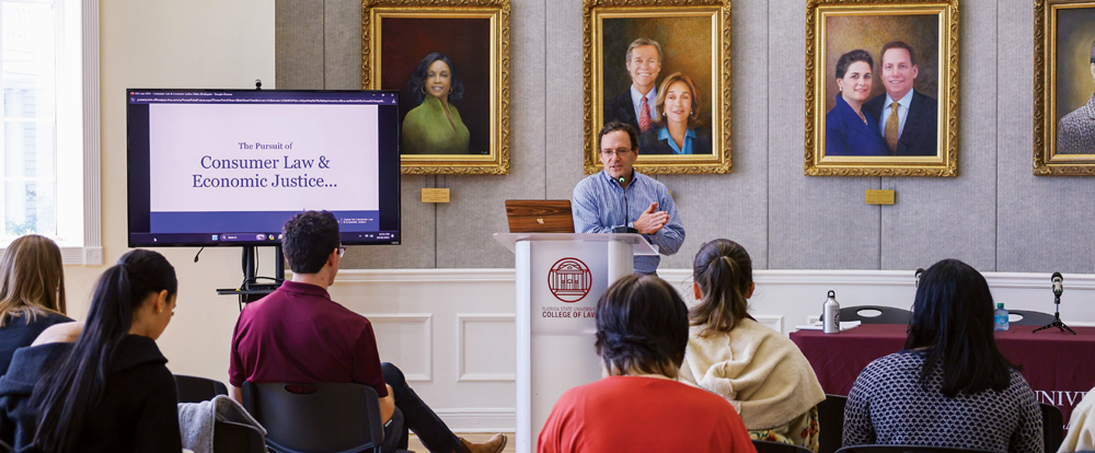 A man speaks at a podium labeled "College of Law," with an audience seated and a presentation slide titled "The Pursuit of Consumer Law & Economic Justice..." displayed on a monitor.