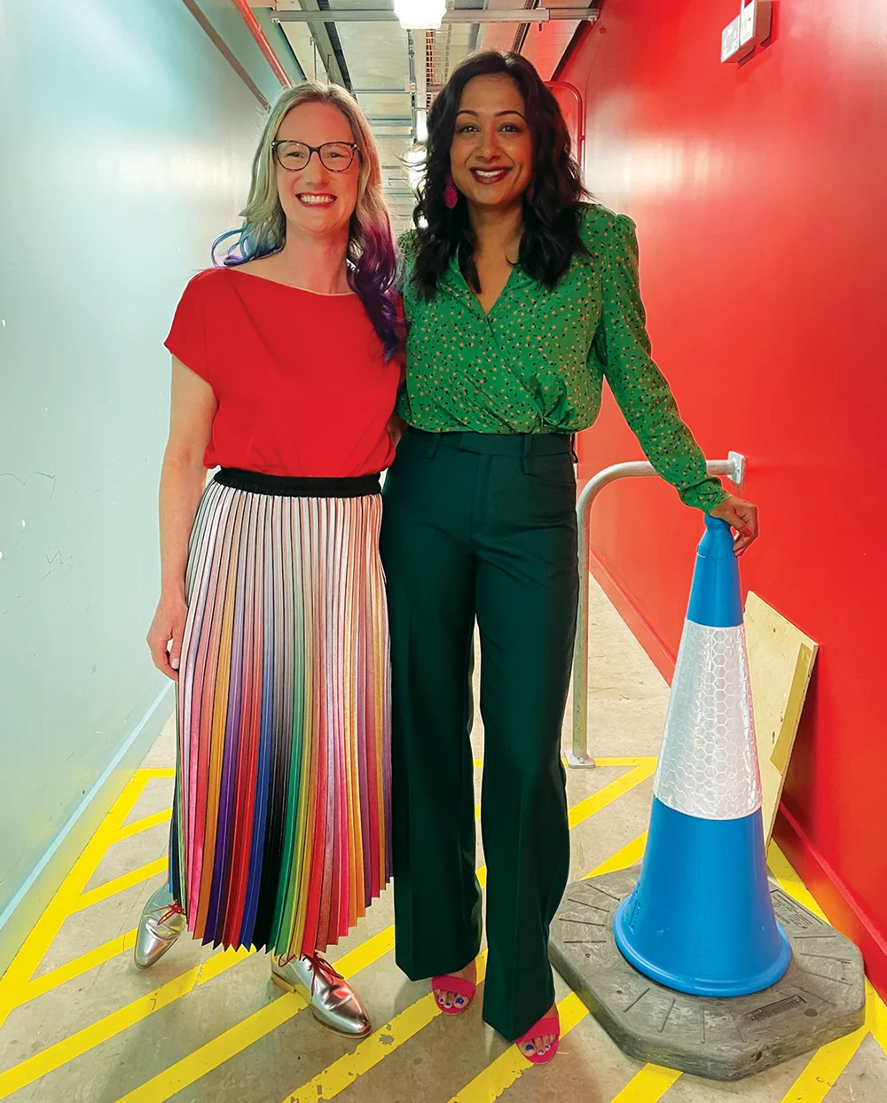 Two people standing in a colorful corridor with a blue and red wall, wearing bright, fashionable clothing, next to a blue traffic cone.