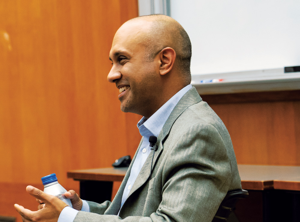 Man in a gray blazer sitting in a chair, holding a water bottle.