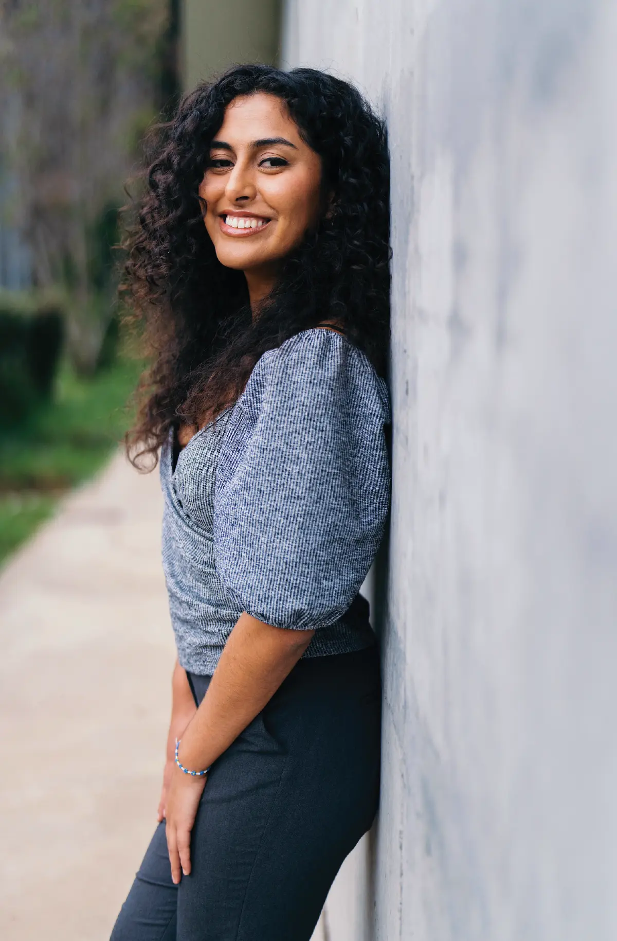 Portrait orientation close-up outdoor photograph view of Krista Arellano smiling and standing posing against the side of a building wall with her hands resting on her dark navy blue dress pants as she is wearing a dark charcoal grey/blue plaid style blouse plus she has a white/blue bead bracelet equipped around her wrist