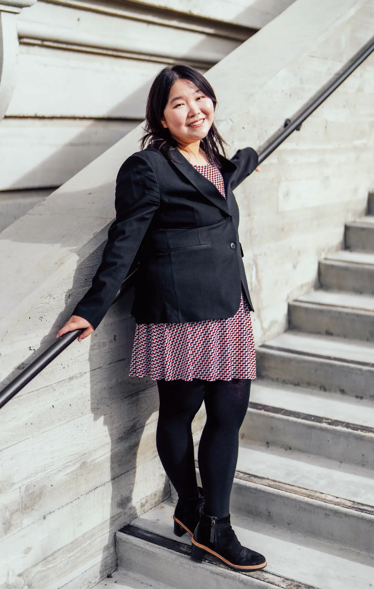 Portrait orientation outdoor close-up photograph view of Tiana Wang smiling and standing on some building stairs nearby a staircase railing as she poses with her arms stretched across the staircase railing while she is wearing a black business blazer suit coat and multi-colored (red/dark navy blue) blouse/dress underneath, black leggings, and black cut heel boot shoes