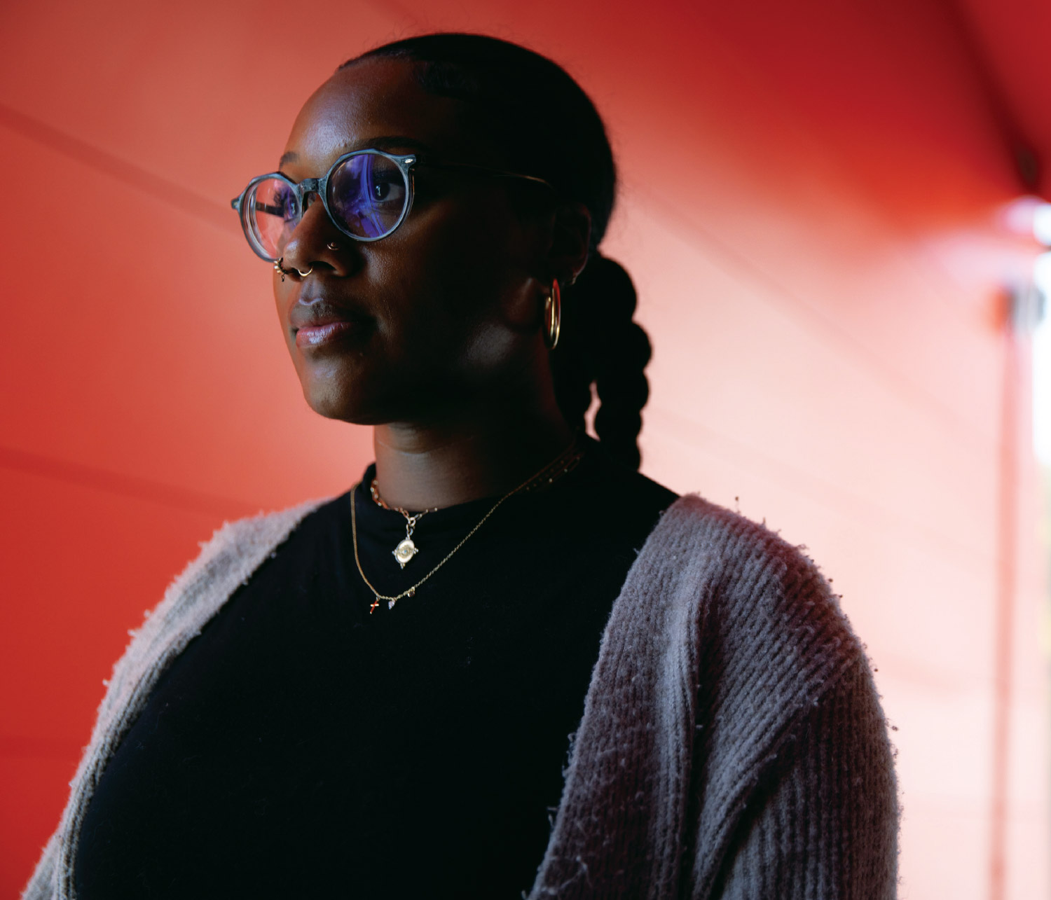 Kaela Allen looking to the left in front of a red orange wall while wearing a black shirt, grey cardigan, and gold piercings and jewelry
